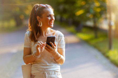 Téléchargez les photos : L'heure d'été. femme élégante souriante en chemise blanche avec sac fourre-tout en utilisant un smartphone et en marchant dans le parc de la ville. - en image libre de droit