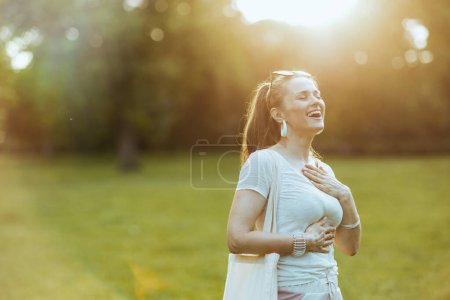 Téléchargez les photos : L'heure d'été. heureuse femme tendance en chemise blanche avec la main sur la poitrine dans la prairie dans le parc de la ville. - en image libre de droit