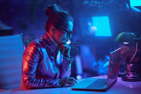 Téléchargez les photos : Neon metaverse concept futuriste. femme élégante dans des lunettes à l'aide d'un ordinateur portable et en utilisant le chat vidéo dans le bureau moderne. - en image libre de droit
