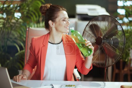 Téléchargez les photos : Un lieu de travail durable. heureuse femme d'affaires moderne au travail en veste rouge avec bouteille d'eau et ventilateur électrique. - en image libre de droit