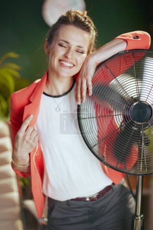 Foto de Lugar de trabajo sostenible. sonriente empleada femenina moderna en moderna oficina verde en chaqueta roja con ventilador eléctrico. - Imagen libre de derechos