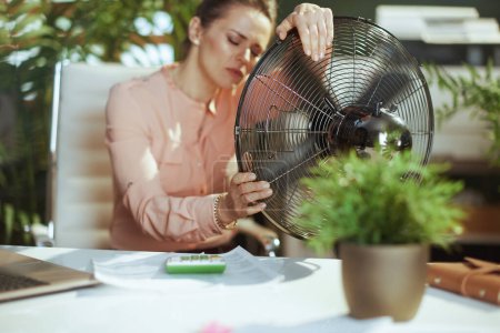 Foto de Lugar de trabajo sostenible. moderna mujer contable de mediana edad en el trabajo con ventilador eléctrico que sufre de calor de verano. - Imagen libre de derechos