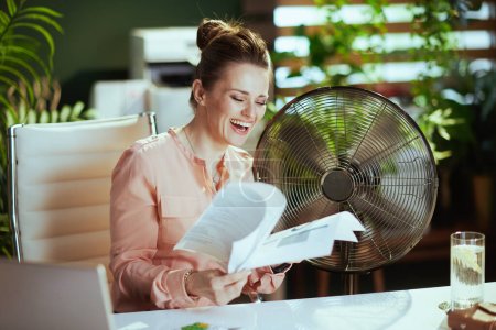 Photo for Sustainable workplace. happy modern woman worker at work with documents and electric fan. - Royalty Free Image