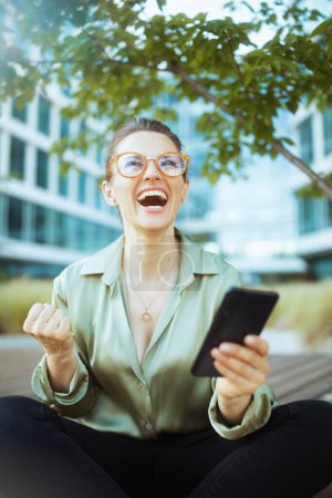 Photo for Smiling modern 40 years old woman employee near business center in green blouse and eyeglasses with smartphone. - Royalty Free Image