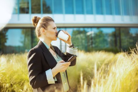 Foto de Mujer trabajadora moderna cerca del edificio de oficinas en chaqueta negra con taza de café usando teléfono inteligente. - Imagen libre de derechos