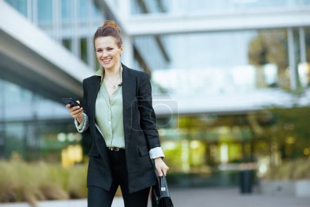 Téléchargez les photos : Souriant moderne femme d'âge moyen travailleur près du centre d'affaires en veste noire avec mallette en utilisant smartphone et la marche. - en image libre de droit