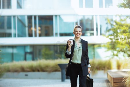 Foto de Feliz mujer trabajadora moderna en el distrito de negocios en chaqueta negra con taza de café caminando. - Imagen libre de derechos