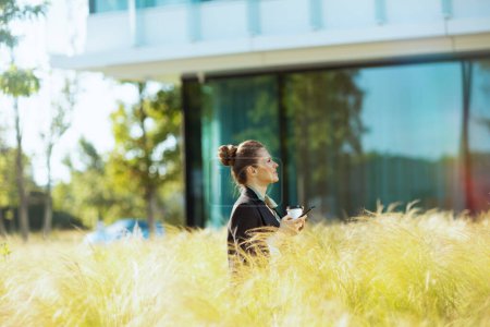 Téléchargez les photos : Chère femme d'affaires moderne près de l'immeuble de bureaux en veste noire avec smartphone et tasse de café. - en image libre de droit