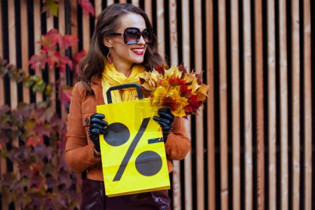 Foto de Hola octubre. Sonriente hembra moderna en gabardina naranja con bolsa de compras y hojas amarillas otoñales en la ciudad. - Imagen libre de derechos