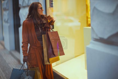 Foto de Hola octubre. mujer moderna sonriente en gabardina marrón con bolsas de compras y hojas amarillas de otoño cerca de la tienda en la ciudad. - Imagen libre de derechos