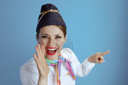 Téléchargez les photos : Heureuse hôtesse de l'air moderne sur fond bleu en uniforme annonçant de bonnes nouvelles pointant quelque chose. - en image libre de droit
