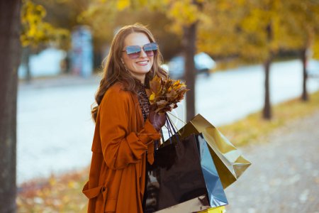 Téléchargez les photos : Bonjour, novembre. Femme élégante et souriante en trench-coat orange avec des sacs à provisions et des feuilles jaune automne dans la ville. - en image libre de droit