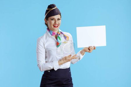Photo for Happy elegant air hostess woman on blue background in uniform showing blank a4 paper sheet. - Royalty Free Image