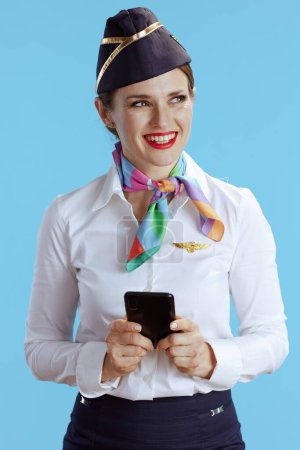 Photo for Happy stylish female stewardess on blue background in uniform sending text message using smartphone. - Royalty Free Image