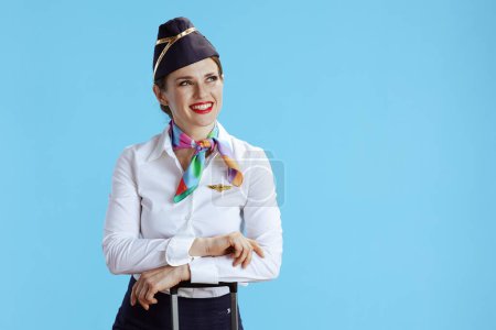 Foto de Sonriente moderna azafata de aire femenino sobre fondo azul en uniforme con bolsa de viaje mirando espacio de copia. - Imagen libre de derechos