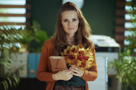 Foto de Joven 40 años de edad propietaria de una pequeña empresa mujer con cuaderno y hojas amarillas de otoño en la moderna oficina verde. - Imagen libre de derechos