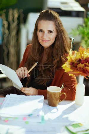 Photo for Smiling young small business owner woman with autumn yellow leaves working with documents in the modern green office. - Royalty Free Image