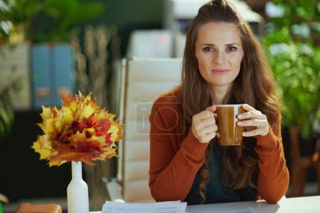 Téléchargez les photos : Femme propriétaire de petite entreprise d'âge moyen avec des feuilles jaunes d'automne et une tasse de thé dans le bureau vert moderne. - en image libre de droit