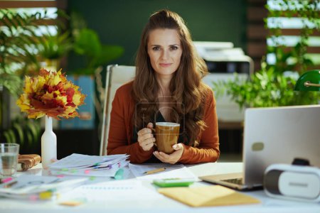 Foto de Feliz elegante 40 años de edad dueño de una pequeña empresa mujer con hojas amarillas de otoño y taza de té en la moderna oficina verde. - Imagen libre de derechos