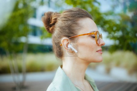 Téléchargez les photos : Employée moderne détendue près du centre d'affaires en chemisier vert et lunettes avec écouteurs sans fil. - en image libre de droit
