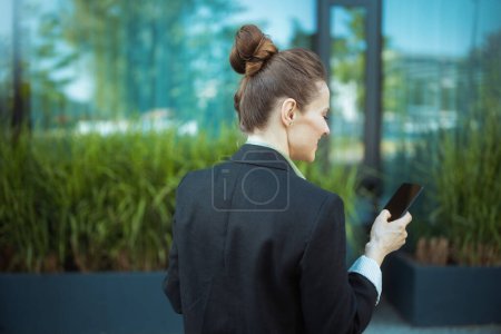 Photo for Seen from behind woman employee near office building in black jacket with smartphone. - Royalty Free Image