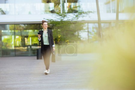 Foto de Retrato de larga duración de una trabajadora moderna de mediana edad cerca del edificio de oficinas en chaqueta negra con maletín usando teléfono inteligente y caminando. - Imagen libre de derechos