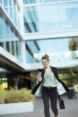 Téléchargez les photos : Heureuse employée moderne dans le quartier des affaires en veste noire avec mallette en utilisant smartphone et marche. - en image libre de droit