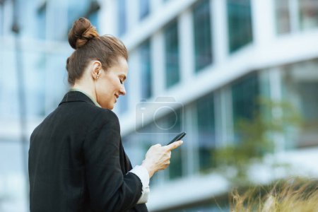 Téléchargez les photos : Vu de derrière femme d'affaires moderne près de l'immeuble de bureaux en veste noire à l'aide d'un smartphone. - en image libre de droit