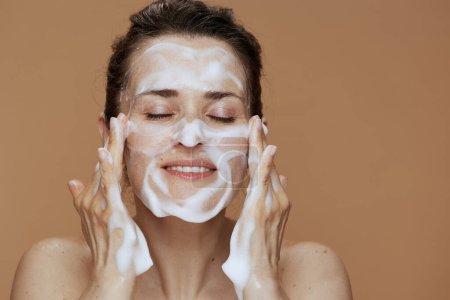 Foto de Feliz joven hembra con rostro espumoso lavar la cara sobre fondo beige. - Imagen libre de derechos