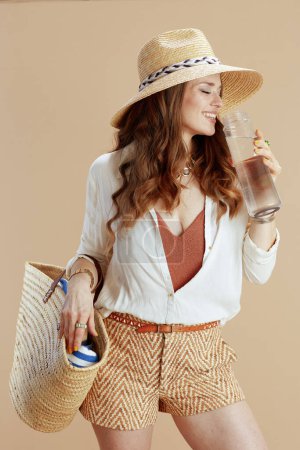 Téléchargez les photos : Vacances à la plage. heureuse femme élégante en chemisier blanc et short sur fond beige avec bouteille d'eau, sac de paille et chapeau d'été. - en image libre de droit
