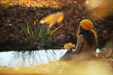 Téléchargez les photos : Bonjour septembre. Vu de derrière femme élégante en manteau beige et chapeau orange avec des feuilles jaune automne assis à l'extérieur sur le parc de la ville en automne. - en image libre de droit