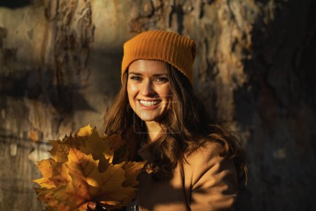 Téléchargez les photos : Bonjour, novembre. Femme élégante souriante en manteau marron et chapeau jaune avec des feuilles jaunes d'automne contre un énorme arbre à l'extérieur dans le parc de la ville en automne. - en image libre de droit
