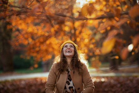 Téléchargez les photos : Bonjour, novembre. souriant femme d'âge moyen à la mode en manteau beige et chapeau orange vomit feuilles d'automne à l'extérieur dans le parc de la ville en automne. - en image libre de droit