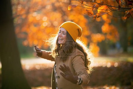 Photo for Hello november. smiling stylish woman in brown coat and yellow hat dancing outside on the city park in autumn. - Royalty Free Image