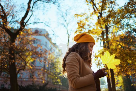 Foto de Hola septiembre. elegante y pensativa mujer de 40 años con abrigo beige y sombrero naranja con hojas amarillas de otoño en el exterior de la ciudad en otoño. - Imagen libre de derechos