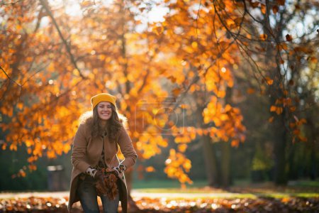 Photo for Hello september. smiling elegant female in beige coat and orange hat throws up autumn leaves outdoors in the city park in autumn. - Royalty Free Image