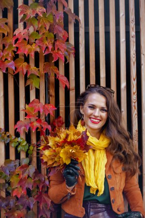 Foto de Hola octubre. hembra sonriente de moda en gabardina naranja con hojas amarillas otoñales en la ciudad. - Imagen libre de derechos