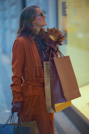Foto de Hola septiembre. sonriente elegante mujer de 40 años en gabardina marrón con bolsas de compras y hojas amarillas de otoño cerca de la tienda en la ciudad. - Imagen libre de derechos