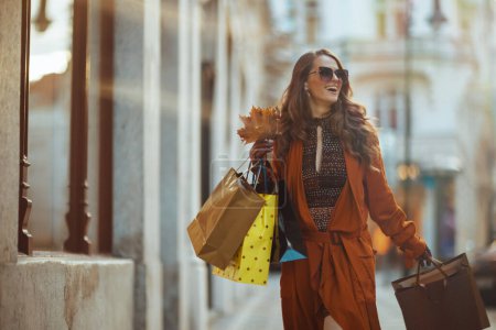 Téléchargez les photos : Bonjour, novembre. heureuse femme d'âge moyen à la mode en trench-coat brun avec des sacs à provisions et des feuilles jaunes d'automne dans la ville. - en image libre de droit