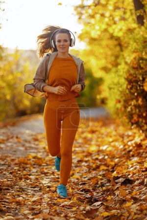 Foto de Hola otoño. Retrato completo de mujer con estilo en ropa de fitness en el parque escuchando la música con auriculares y jogging. - Imagen libre de derechos