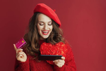 Foto de Feliz San Valentín. sonriente elegante mujer de mediana edad en suéter rojo y boina con corazón rojo, teléfono inteligente y tarjeta de crédito navegación tienda en línea. - Imagen libre de derechos