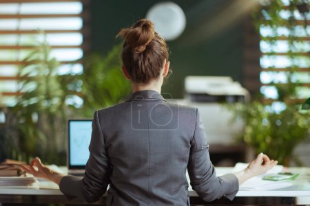 Photo for Sustainable workplace. Seen from behind modern small business owner woman in a grey business suit at work meditating. - Royalty Free Image