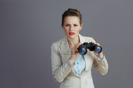 Téléchargez les photos : Élégante femme d'affaires d'âge moyen en costume d'affaires léger avec jumelles isolées sur fond gris. - en image libre de droit
