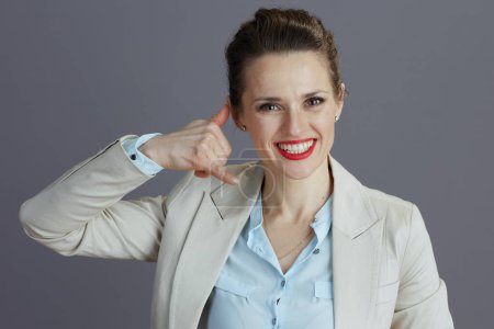 Foto de Retrato de una empleada elegante sonriente en un traje de negocios ligero que muestra llámame gesto contra fondo gris. - Imagen libre de derechos