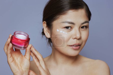 Foto de Modern asian female with facial cream jar and eye cream on face against blue background. - Imagen libre de derechos