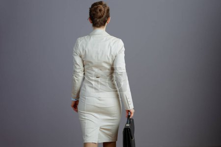 Foto de Visto desde atrás empleado femenino en un traje de negocios ligero con maletín caminando sobre fondo gris. - Imagen libre de derechos