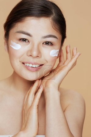 Téléchargez les photos : Portrait de femme asiatique moderne avec crème faciale sur le visage sur fond beige. - en image libre de droit