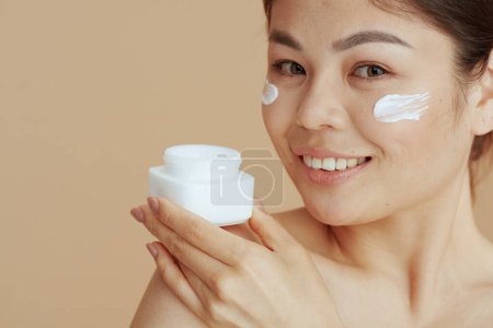 Foto de Retrato de mujer moderna con frasco de crema facial y crema facial en la cara contra fondo beige. - Imagen libre de derechos