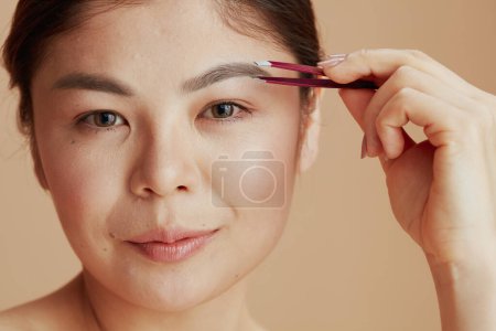Téléchargez les photos : Portrait de jeune femme asiatique avec pince à épiler sur fond beige. - en image libre de droit
