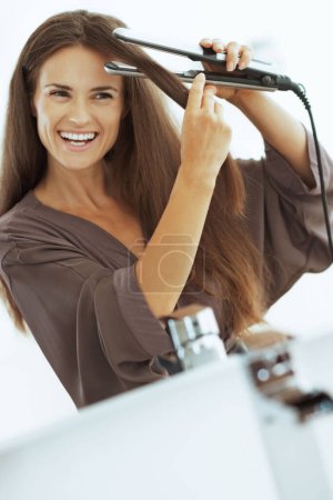 Téléchargez les photos : Souriant jeune femme lissant les cheveux avec lisseur - en image libre de droit